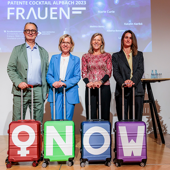 Patente Cocktail 2023 in Alpbach unter dem Motto: Patente Frauen. Auf der Bühne 1 Mann und 3 Frauen mit jeweils einem Koffer. Auf den Koffern sind Aufkleber mit dem Symbol für weibliches Geschlecht und den Buchstaben N, O und W - ergibt das wort now, englisch für jetzt. 