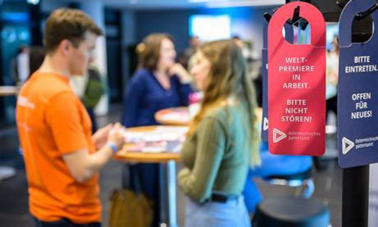 im Vordergrund sind 2 Türschilder mit der Aufschrift "Weltpremiere in Arbeit" und "Offen für Neues" zu sehen. Im Hintergrund sind sich unterhaltende Personen verschwommen zu sehen.