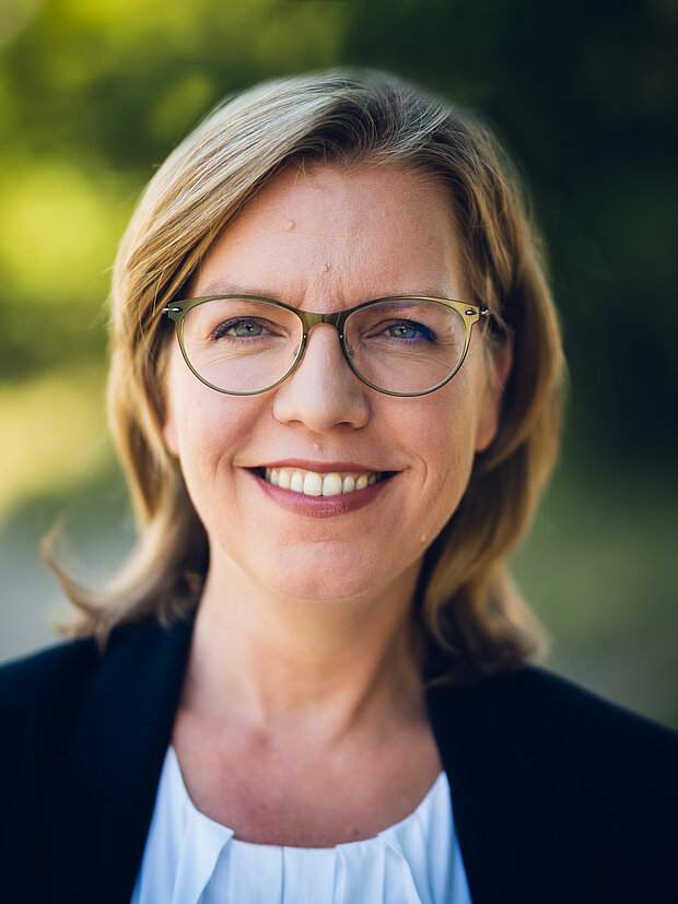 Portrait von Bundesministerin Leonore Gewessler. Sie steht im Freien, im Hintergrund sind verschwommen Bäume erkennbar. 